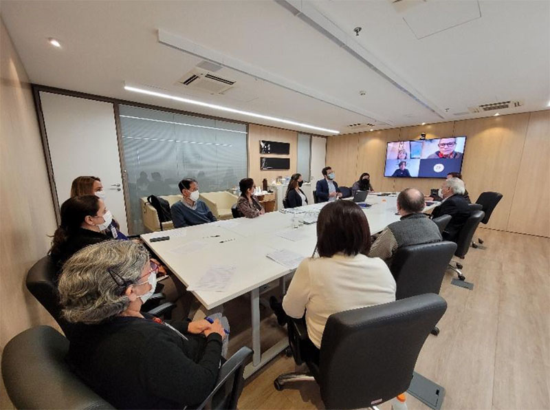 Sala de reunião, com uma grande mesa clara e, ao redor dela, 12 pessoas sentadas em cadeiras. Elas estão participando de uma telechamada: olham em direção a uma grande televisão fixada à parede. Na tela, aparece o rosto de pessoas que estão participando da reunião à distância.