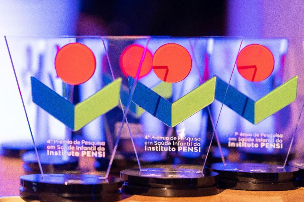 Três troféus do prêmio. Eles têm uma base escura e, sobre ela, uma estrutura vertical transparente, com o logotipo do Instituto PENSI e, abaixo dele, a frase: 4º Prêmio Pesquisa em Saúde Infantil do Instituto PENSI.