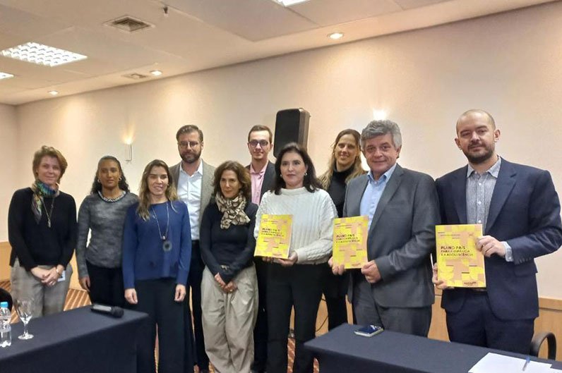 Grupo de dez pessoas (seis mulheres e quatro homens) de costas para uma parede clara e entre duas mesas retangulares cobertas de tecido azul-marinho. No centro do grupo está a então candidata a presidente Simone Tebet, com calça azul-marinho e agasalho branco. Ela e outros dois homens a seu lado (ambos de paletó) estão segurando um livro de capa amarela com o título: “Plano País para a Infância e a Adolescência”.