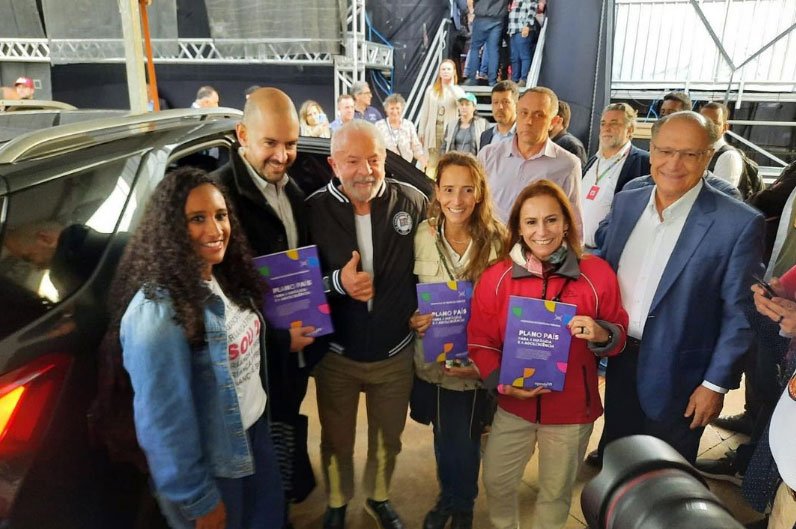 Grupo de seis pessoas (três mulheres e três homens), num ambiente coberto, mas próximas de um carro preto com porta aberta. No centro do grupo está o então candidato a presidente Lula, com agasalho preto semiaberto sobre camiseta branca, calça marrom-clara e tênis preto. Duas mulheres do seu lado esquerdo e um homem de seu lado direito seguram um livro de capa roxa com o título: “Plano País para a Infância e a Adolescência”. A pessoa mais à direita na foto é o então candidato a vice-presidente Geraldo Alckimin, que está de paletó azul aberto e camisa branca de gola.