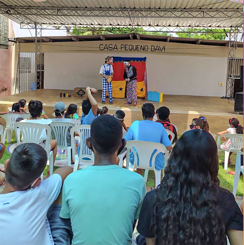 Foto de uma apresentação de dois palhaços. Eles estão num cenário com partes coloridas, que formam um teatro de pequenas dimensões. Ao fundo, há uma parede branca com uma faixa cinza, onde está escrito: Casa Pequeno Davi. Em frente ao palco pode-se ver uma plateia de jovens e adolescentes (a maioria de camiseta azul-claro). Eles estão sentados em cadeiras brancas de plástico.