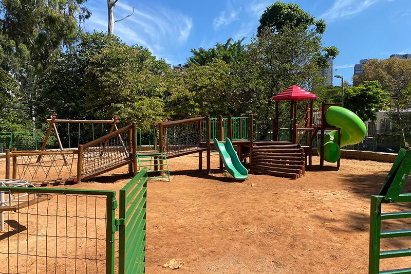 Imagem de um parquinho com chão de areia e cercado por árvores altas. No parquinho pode-se ver um gira-gira e um brinquedo com uma rampa de madeira longa, mas suave, que leva até um escorregador de plástico verde. Continuando na mesma estrutura há mais um trecho de madeira e uma escada com nove degraus de toras cumpridas. No parquinho também há uma estrutura mais alta, que dá para um escorregador maior, também verde, que desce em forma de caracol]