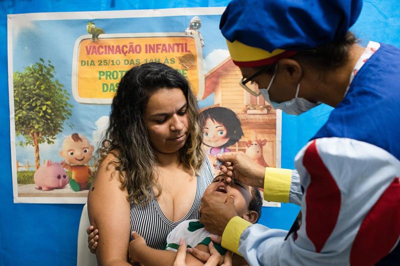 Agente de saúde pinga uma gota na boca de uma criança pequena, que está no colo de uma mulher de cabelos crespos. Ao fundo, há uma parede azul com um grande cartaz onde é possível ler: Vacinação Infantil.