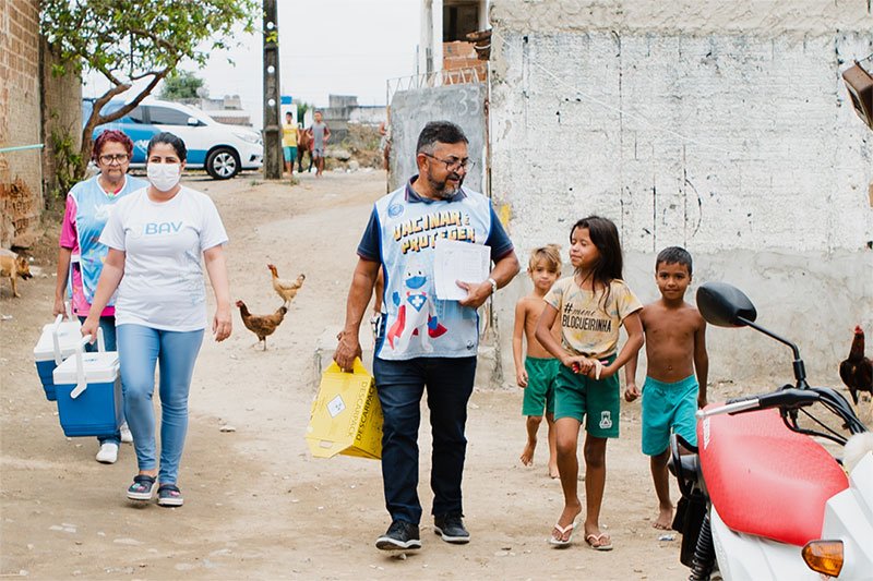 Três técnicos de saúde (duas mulheres e um homem) andando numa rua de terra. Ao lado deles há três crianças. Na camiseta do homem é possível ler: Vacinar protege