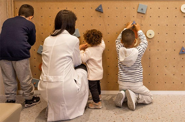 Três crianças brincando junto a uma parede com furos onde se colocam objetos. Uma mulher de avental branco está agachada, próxima às crianças, auxiliando uma delas.