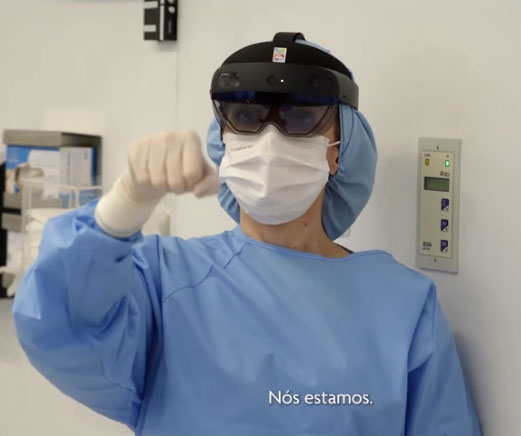 Imagem capturada de vídeo, com uma médica vestida de macacão azul, touca azul, luva branca, óculos de realidade virtual e máscara branca cobrindo nariz e boca.