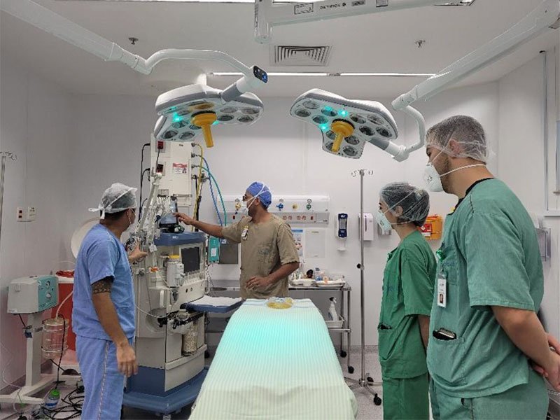 Sala de hospital, com uma cama no meio e equipamentos médicos ao fundo. À esquerda da cama há dois homens, ambos com uniforme, touca e máscara branca. À direita, outras duas pessoas, uma mulher e um homem, também com touca e máscara. Um dos profissionais do lado esquerdo mexe em um grande aparelho, e está sendo observado pelos outros três.