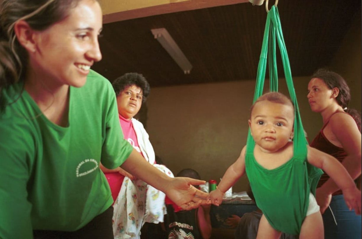 Uma agente de saúde sorridente, de camiseta verde (com logo branco da Pastoral da Criança), pesa um bebê usando uma estrutura de pano verde-escuro. A cena é observada, ao fundo, por uma mulher de camiseta rosa segurando um pano de criança. Em frente dela há outra mulher, com camiseta regata marrom.