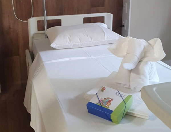 Cama de hospital, coberta com roupa-de-cama toda branca. Uma das peças está dobrada sobre o lençol, em forma de um pequeno elefante.