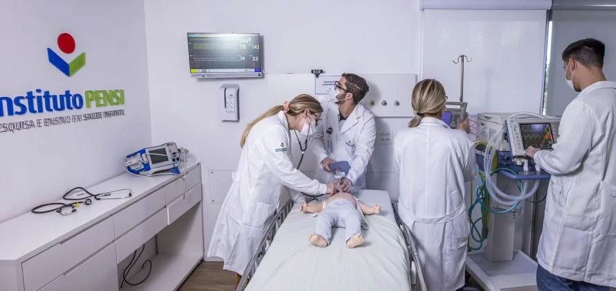 A imagem mostra o interior de uma sala toda branca. Na parede à esquerda, aparece a marca Instituto PENSI e seu logotipo, além de uma bancada com estetoscópios e um pequeno monitor. Na parte central da imagem, há uma cama com lençol branco e, sobre ela, um bebê (um boneco simulador), vestido também de branco. Uma mulher loira, de cabelo preso, máscara médica branca e avental branco, está à esquerda da cama, com uma das mãos próxima à cabeça do boneco. Atrás da cama, há um homem, também de avental branco e máscara médica branca. Ele segura equipamentos médicos, encostados à cabeça do boneco, e olha para um monitor de UTI, posicionado na parede atrás dele. Há ainda mais dois profissionais de jaleco branco na sala, à direita da cama. Os dois (outra mulher loira e um homem de cabelo curto) estão de costas para a foto, olhando outros monitores de acompanhamento do paciente.]