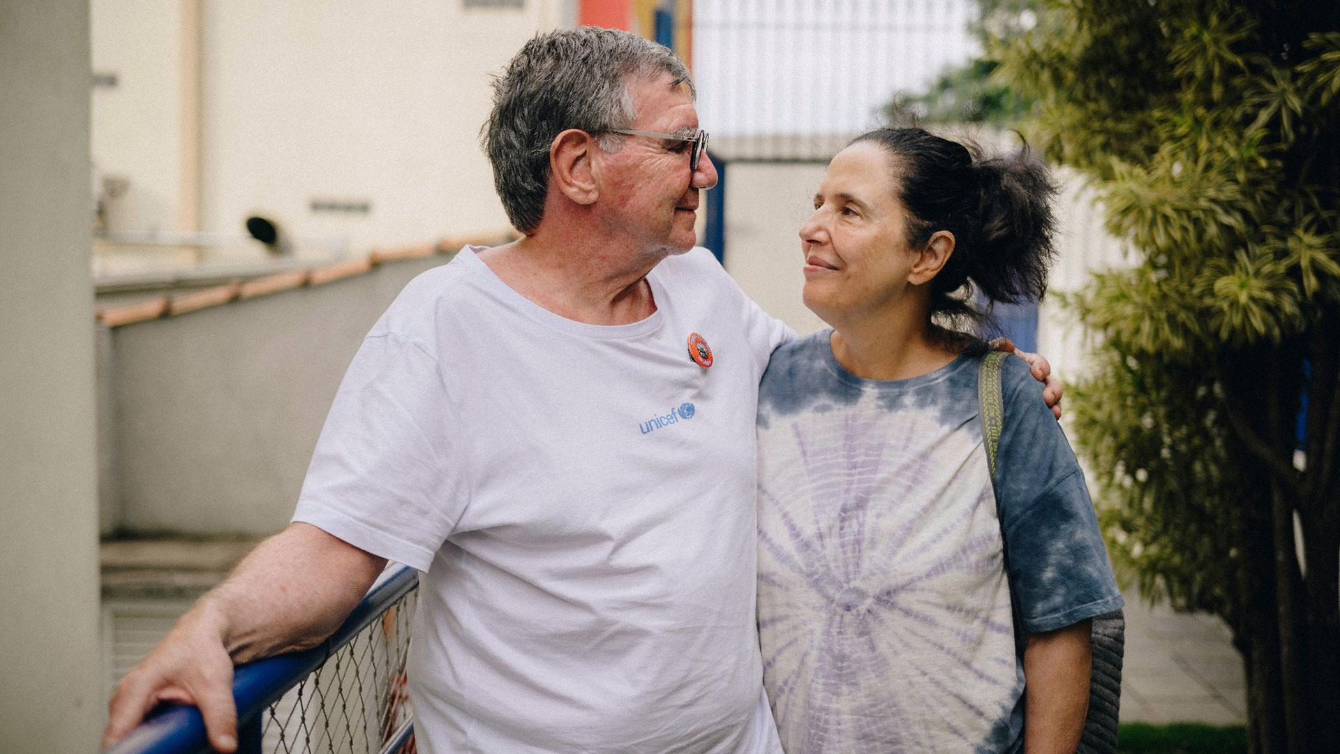 Foto de José Luiz Setúbal e Sandra Mutarelli Setúbal. Eles estão lado a lado, sorrindo, um olhando para o outro. Ele está de camiste branca com o logotipo do Unicef e ela com uma camiseta azul, branca e com leves tons de lilás.