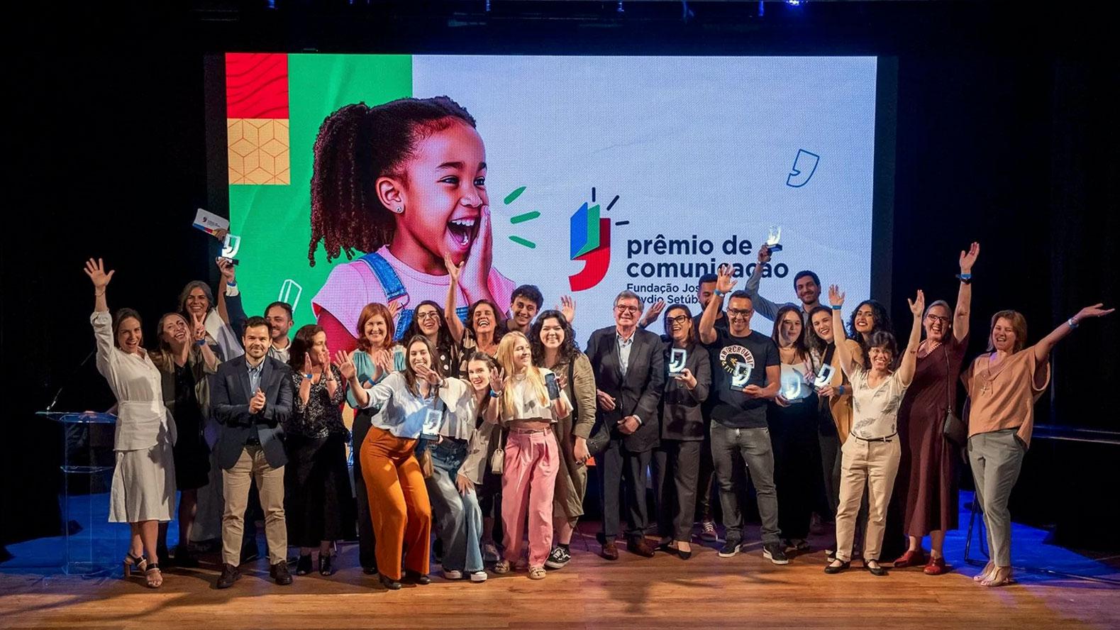 Grupo de homens e mulheres posando para a foto. Vários estão com as mãos para o alto, num gesto de comemoração. Eles seguram o troféu do prêmio. Ao fundo, há uma tela de projeção com o layout do prêmio de comunicação