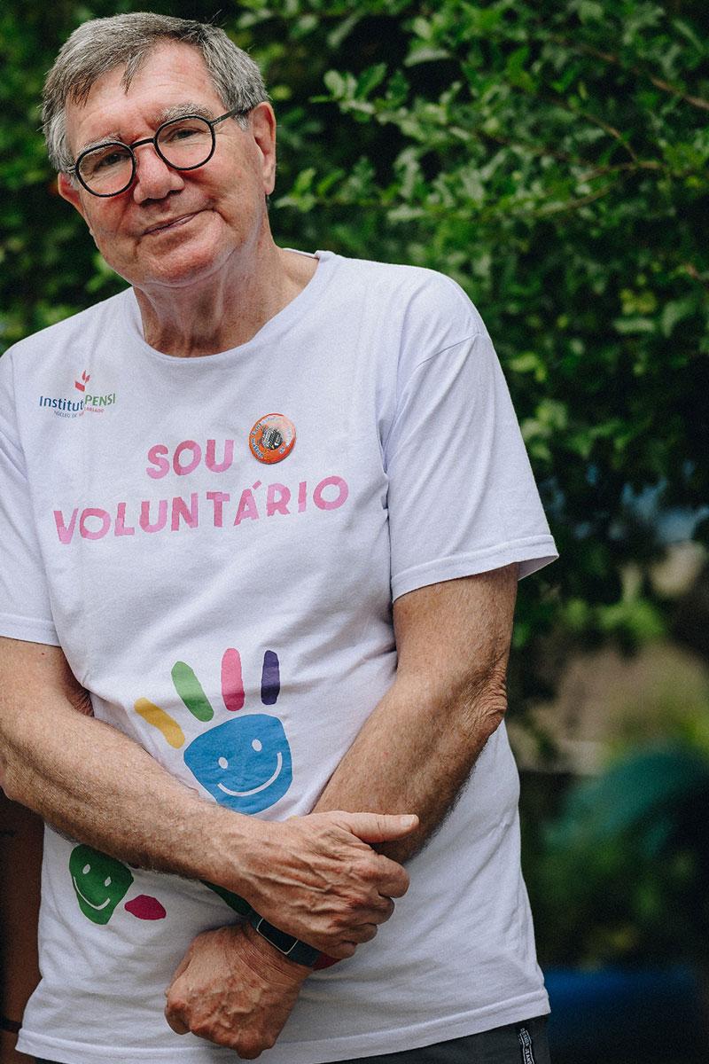 Foto de José Luiz Egydio Setúbal. Ele é um homem grisalho, com óculos de aro preto e uma camiseta branca. A camiseta tem ilustrações infantis e a frase "Sou voluntário"