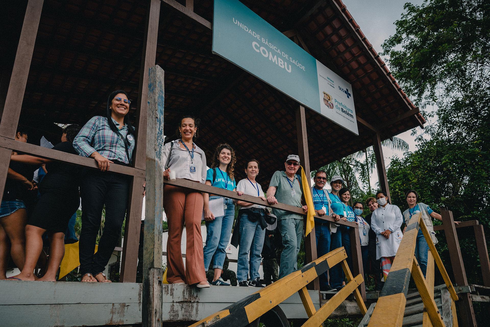 Grupo de pressoas reunidas em uma área semicoberta, tendo acima uma placa que diz "Unidade Básica de Saúde Combu". O grupo é composto por homens e mulheres de diferentes idades, todos sorrindo para a câmera. A maioria está usando calças e camisetas casuais