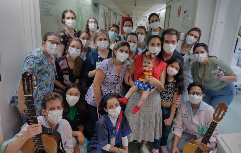 Grupo de homens e, principalmente, mulheres posando para a foto. Todos estão com máscara facial. Em primeiro plano, há um homem e uma mulher agachados, cada um segurando um violão. No centro da foto está a única pessoa sem máscara: uma bebê com roupinha de mulher maravilha