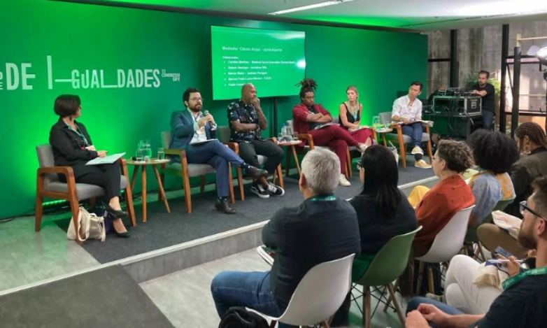 Foto da plenária, com seis pessoas sentadas em cadeiras no palco e parte da plateia assistindo