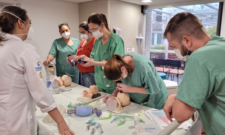 A foto mostra seis pessoas — três delas de jaleco verde, uma de jaleco branco do Sabará. Elas estão ao redor de uma mesa com diversos equipamentos e bonecos médicos