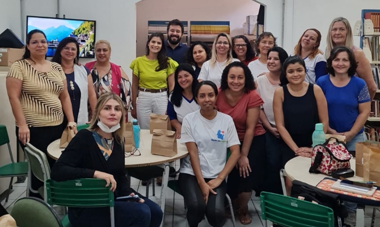 Um homem e diversas mulheres posando para a foto, num ambiente fechado com duas mesas redondas, algumas cadeiras e uma tela de TV ao fundo