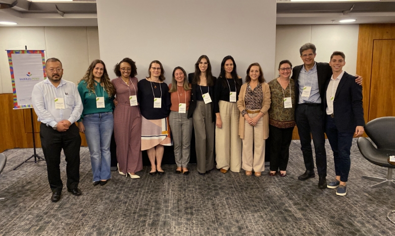 Diversas pessoas, a maioria mulheres, posando para a foto num ambiente com carpete cinza e branco e uma tela de projeção ao fundo. Elas estão vestidas de modo um pouco formal e usam crachá