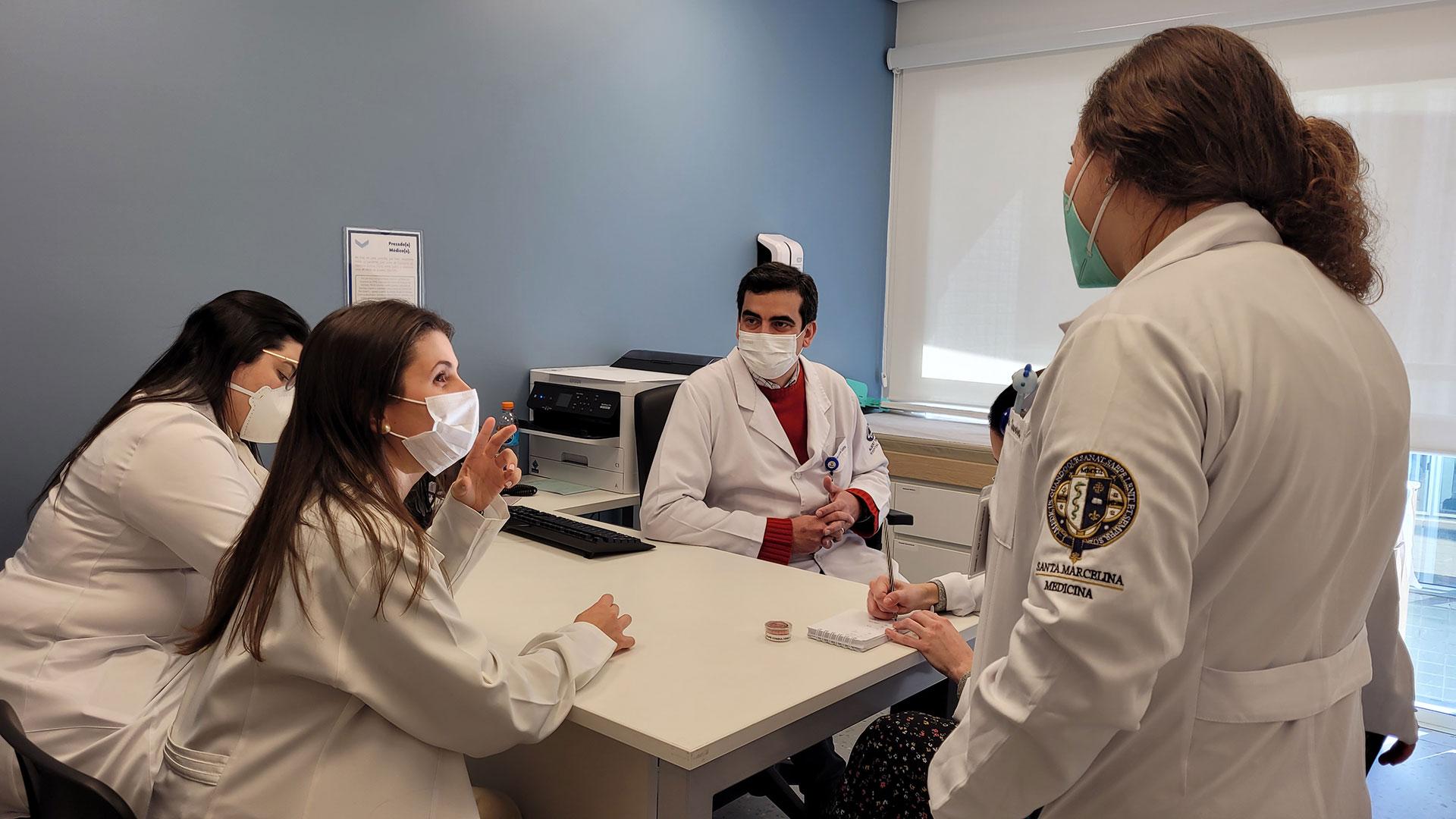 Pequeno conjunto de pessoas de jaleco branco e máscara facial branca, ao redor de uma mesa branca