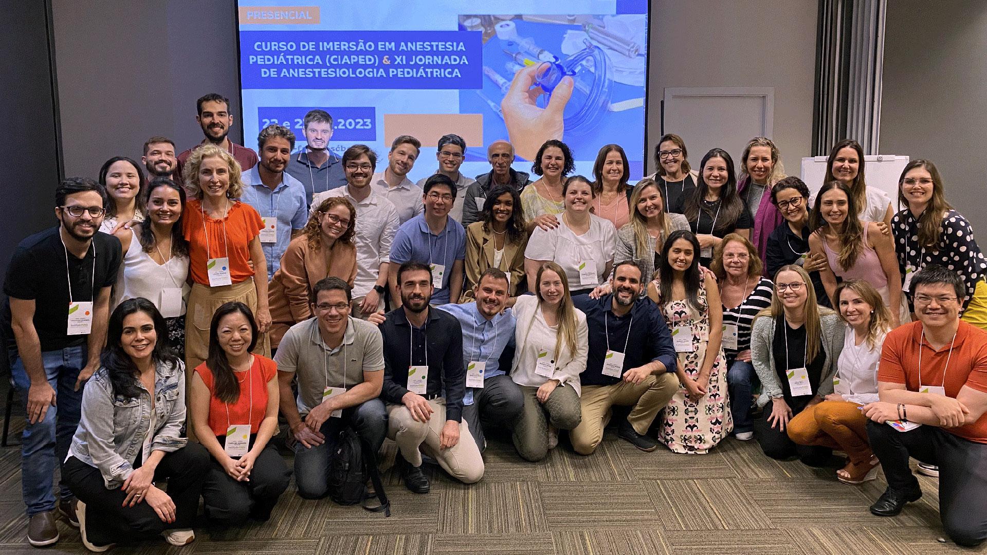 Grupo de homens e mulheres posando para a foto. Ao fundo, há um telão com o nome de um curso de anestesia pediátrica