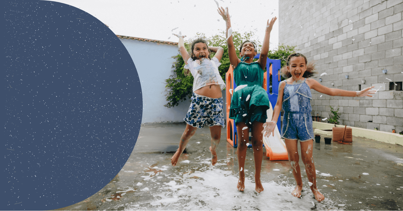 Três meninas em uma área aberta, brincando com bolhas de sabão. Elas estão uma ao lado da outra, com expressões felizes e sorridentes. As meninas usam roupas casuais e coloridas