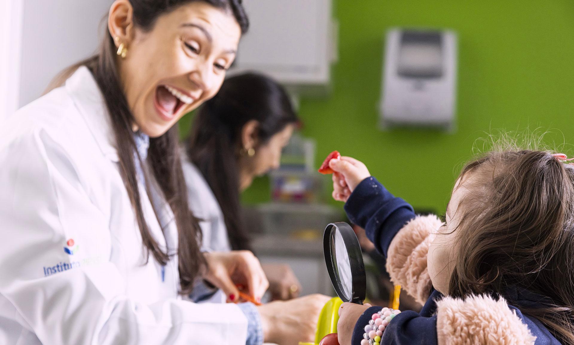 Uma mulher de jaleco branco, com a boca bem aberta, está sorrindo para uma menina que lhe estende a mão direita com um pedaço de comida