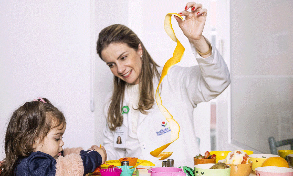 Uma mulher de cabelo claro, vestindo jaleco branco do Instituto Pensi, está com a mão erguida, segurando uma comprida casca de laranja. A seu lado está uma garotinha de cabelo castanho. As duas estão à frente de uma mesa com vários potes com frutas